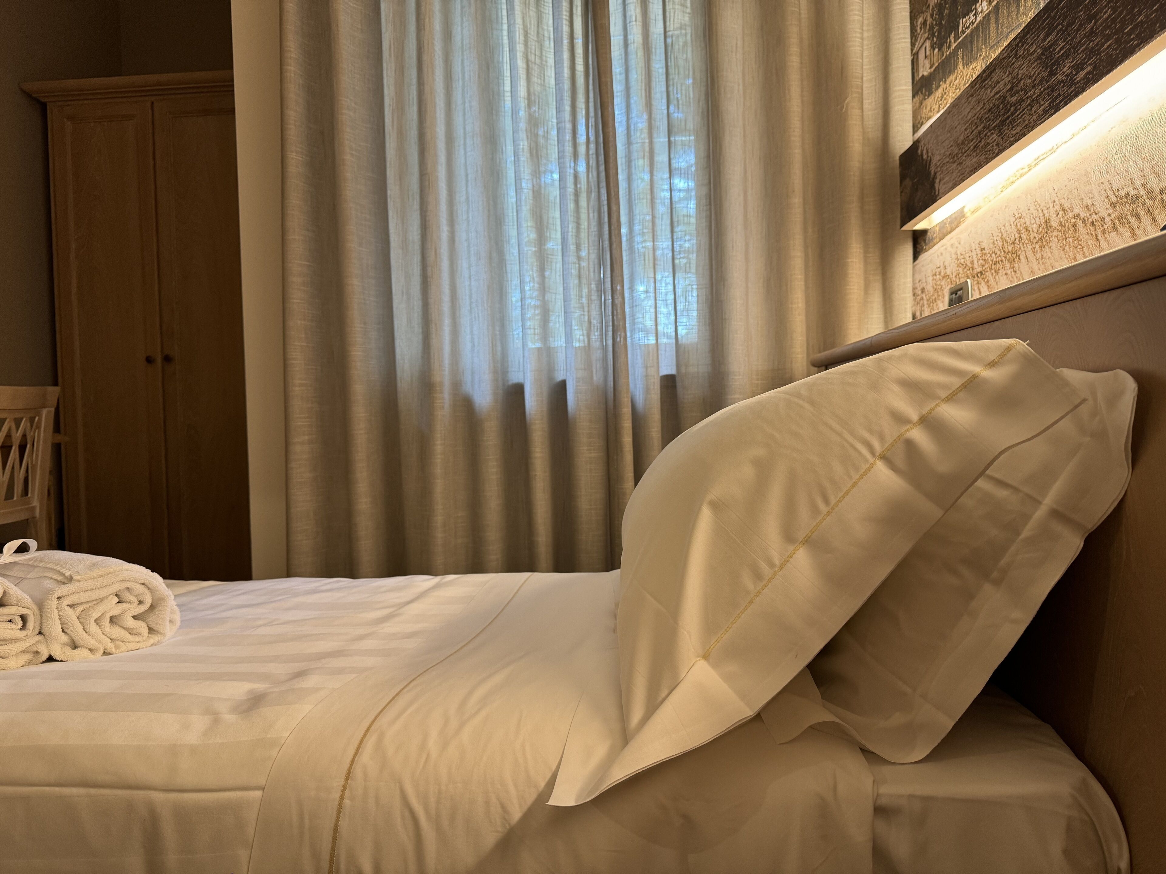 A neatly made bed with a pillow and rolled towels, a wooden wardrobe, and a window with curtains.