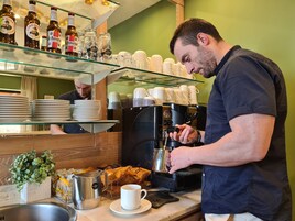 Un barista preparando café en una cafetería con estantes llenos de botellas de cerveza, pilas de platos y una máquina de café.