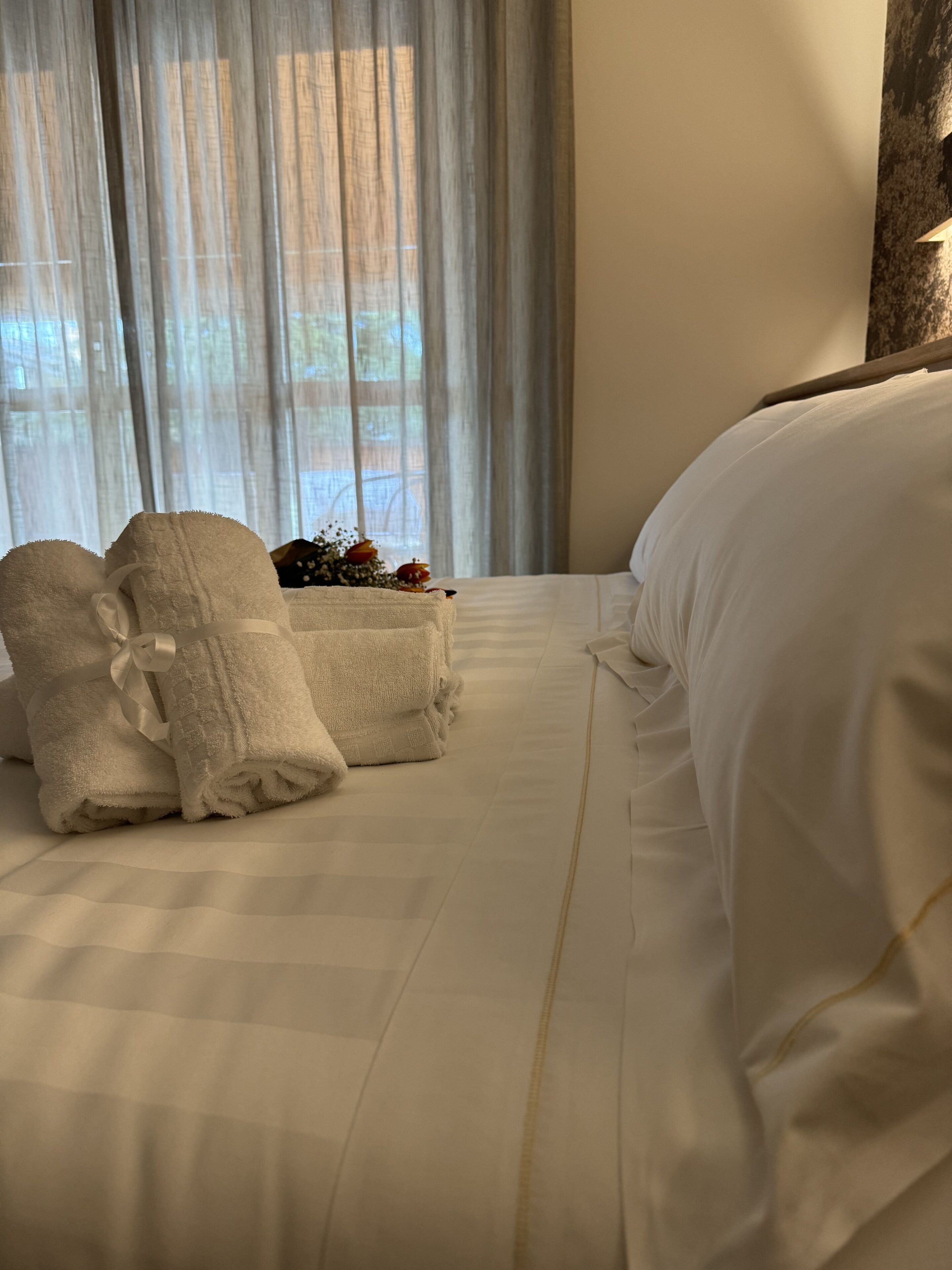 Ein ordentlich gemachtes Bett mit weißen Leinen und zwei gefalteten Handtüchern mit Schleife, ein kleines dekoratives Element auf dem Bett und im Hintergrund transparente Vorhänge.