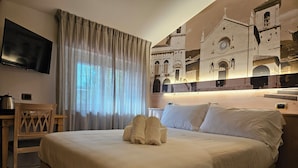 Habitación de hotel con una cama grande, un televisor de pantalla plana en la pared, una silla de madera, un escritorio con un hervidor y vistas a un edificio histórico a través de cortinas translúcidas.