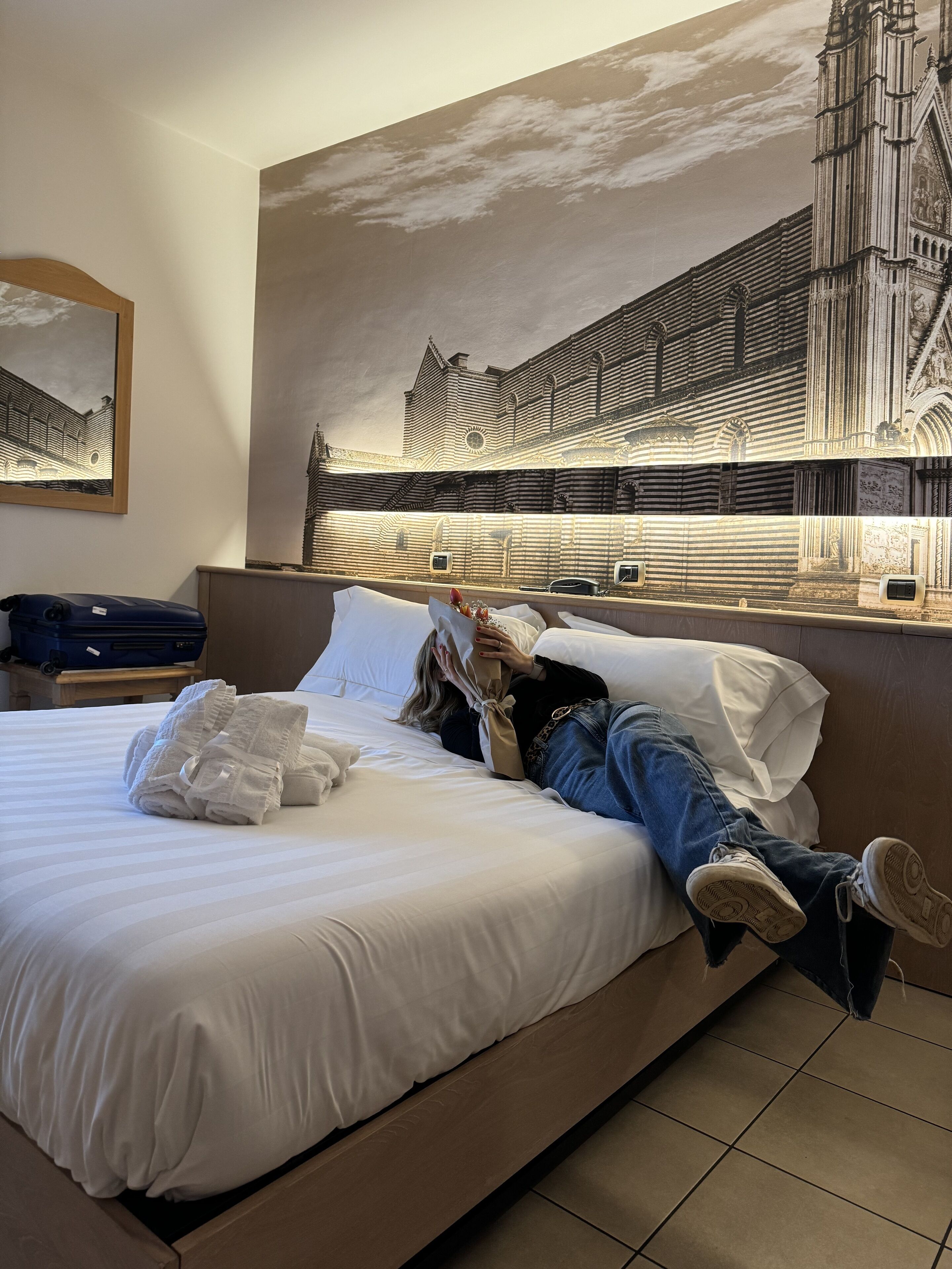 Habitación de hotel con una cama grande, un cuadro de un edificio, una maleta y una persona acostada en la cama.
