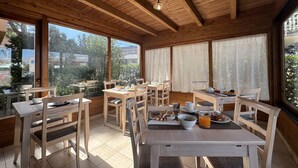Un área de desayuno con mesas y sillas de madera, ventanas amplias con cortinas y vistas al exterior.