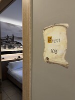 Una habitación con una cama, un letrero en la pared que dice "Revi 103" y vistas a un pueblo con una iglesia y montañas al fondo.