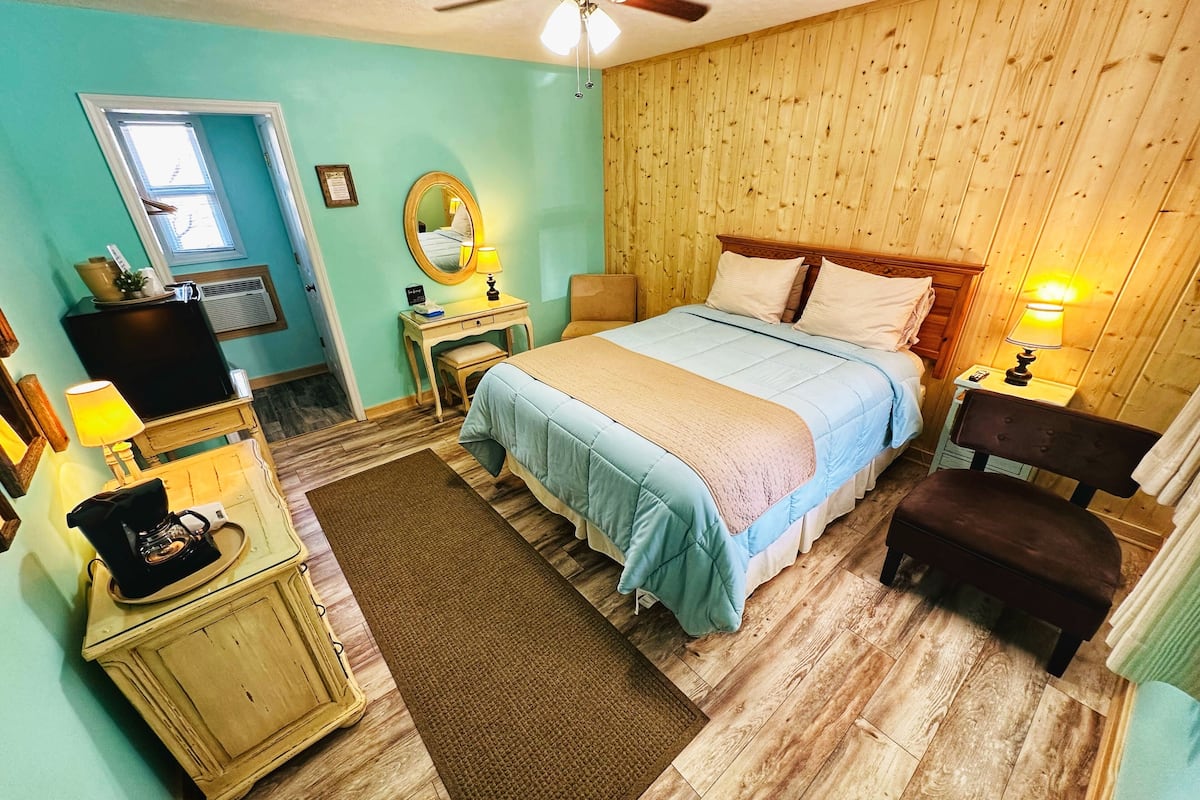 Un dormitorio con cama, escritorio, silla, espejo, ventilador de techo y ventana.