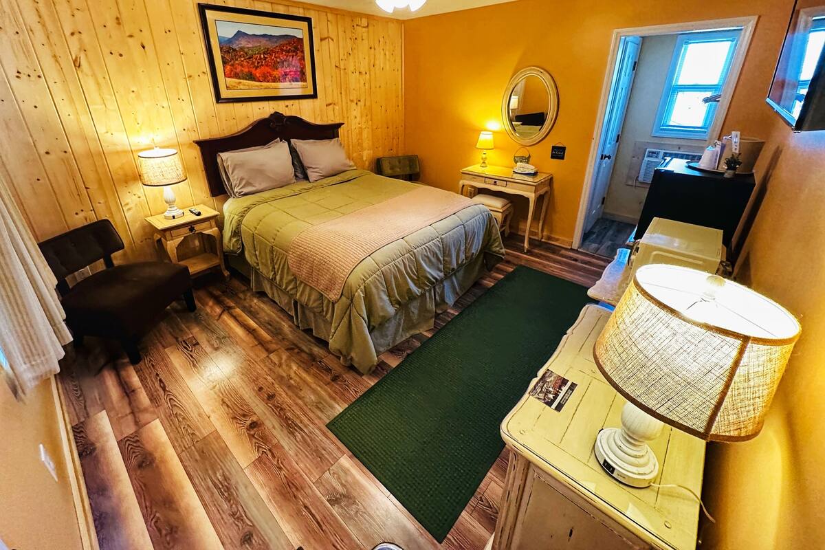 Un dormitorio acogedor con piso de madera, cama con colcha verde, mesita de noche con lámpara, cómoda con lámpara y ventilador de techo.