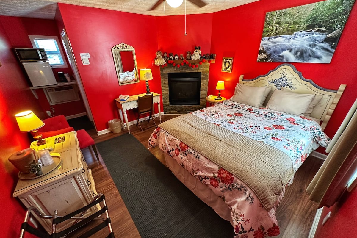 Un dormitorio con una pared roja, una cama con ropa de cama floral, un escritorio con lámpara, un espejo y una chimenea.