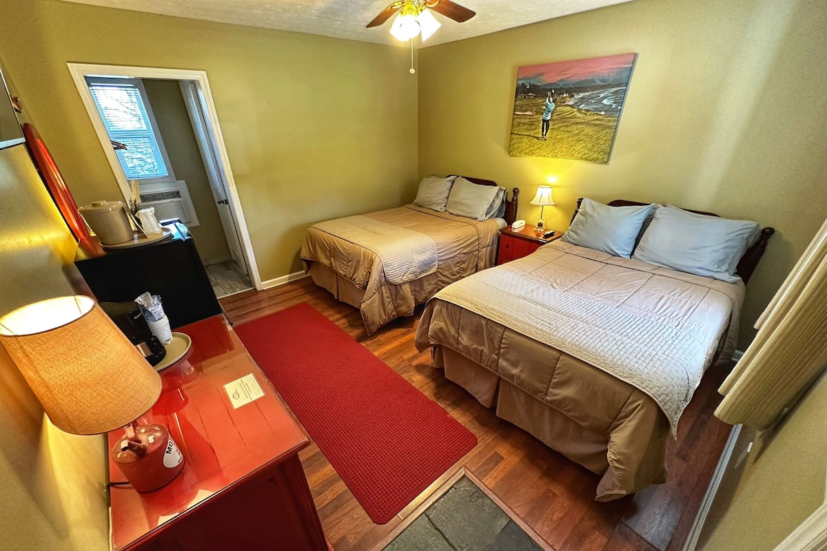 Habitación de hotel con dos camas, un escritorio con lámpara, un tapete rojo y un cuadro en la pared.