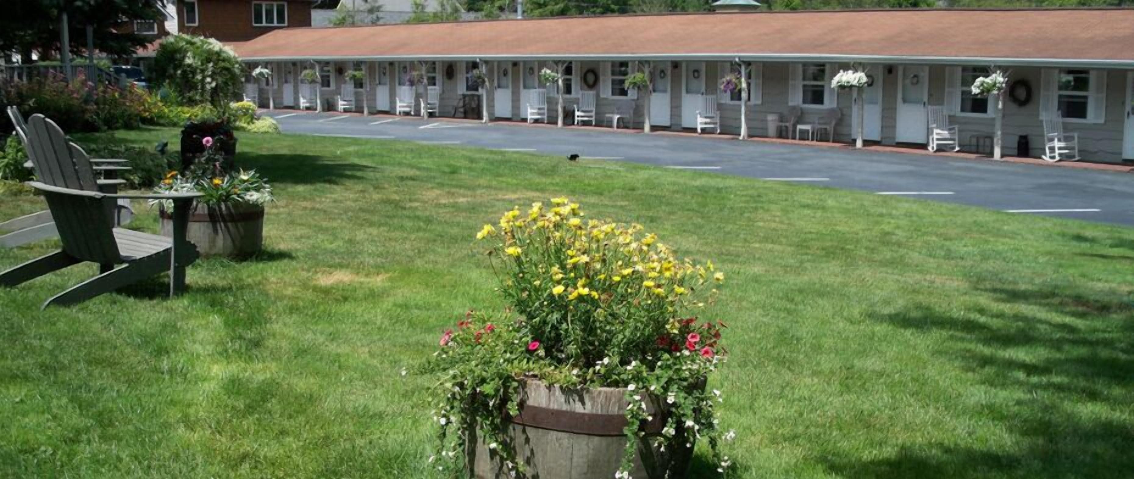 A motel with a row of single-story rooms, a covered parking area, and a grassy lawn with flower beds.