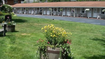 A motel with a row of single-story rooms, a covered parking area, and a grassy lawn with flower beds.