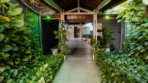 A covered walkway flanked by lush green plants, leading to a room with a wooden ceiling and a view of the interior. - Hotel Village Premium Caruaru (Caruaru)