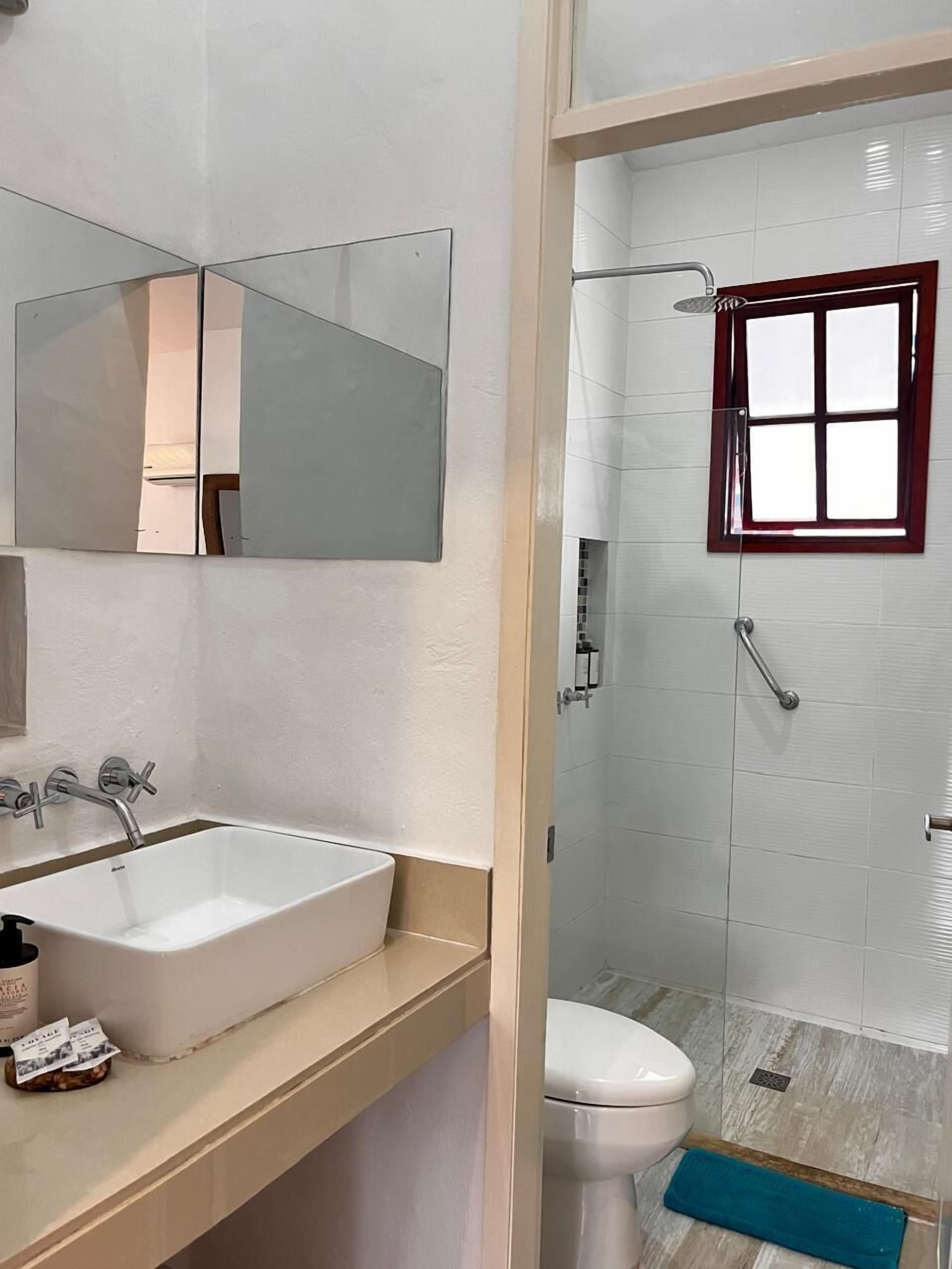 Un baño con lavamanos blanco, espejo y zona de ducha con ventana.