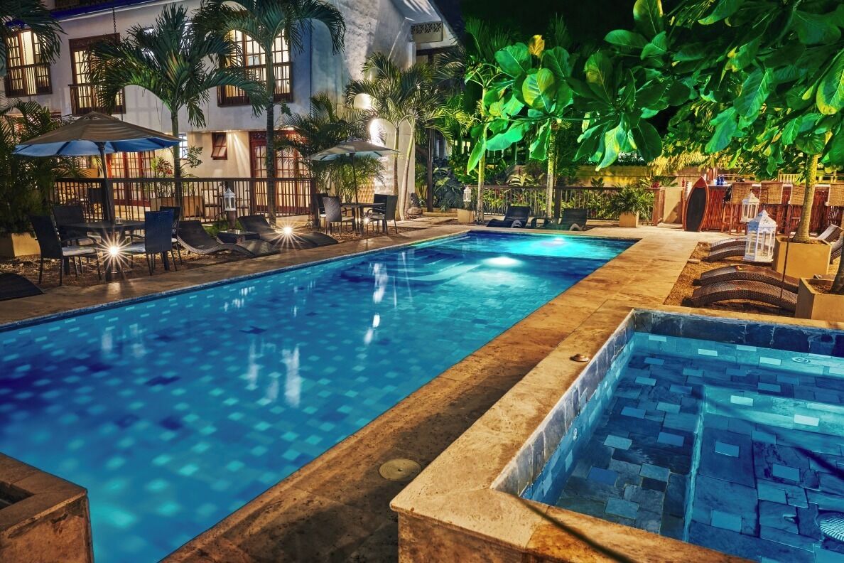 Una piscina con sillones de descanso y sombrillas, rodeada de palmeras y un edificio con grandes ventanales.