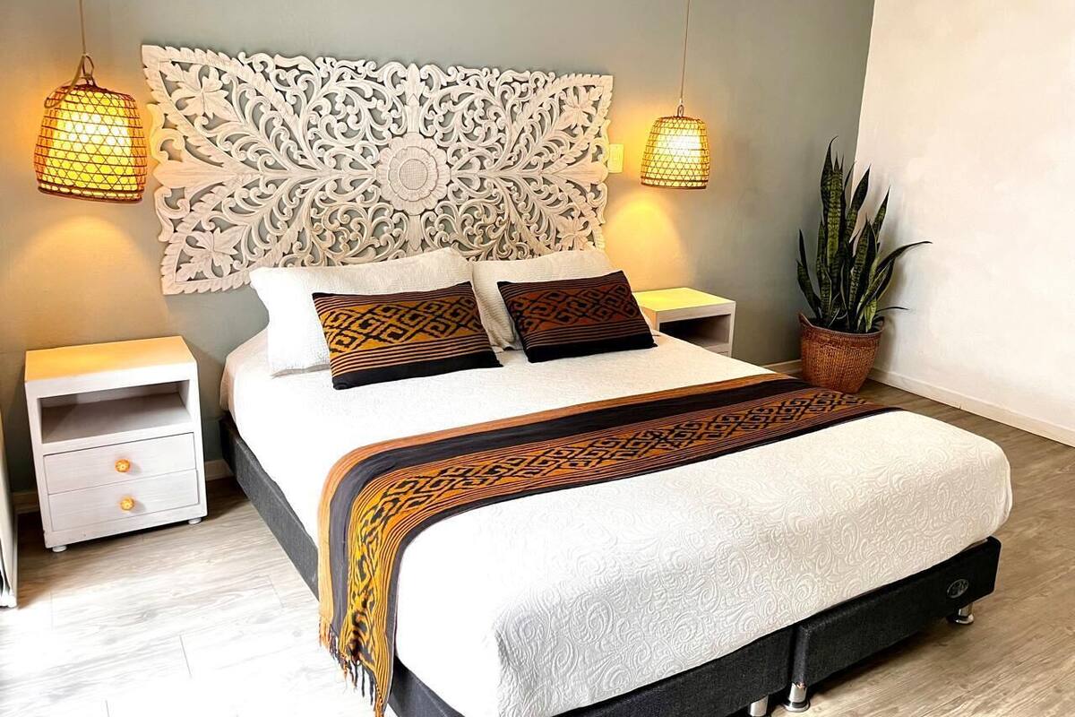 Une chambre à coucher moderne avec un grand lit, deux tables de chevet, une tête de lit décorative et deux lampes suspendues.