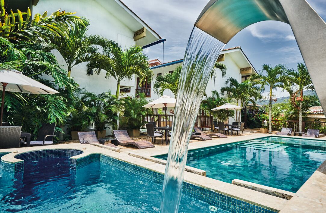 Ein Pool mit Wasserfall, umgeben von Liegestühlen und Sonnenschirmen, mit Palmen und einem Gebäude im Hintergrund.