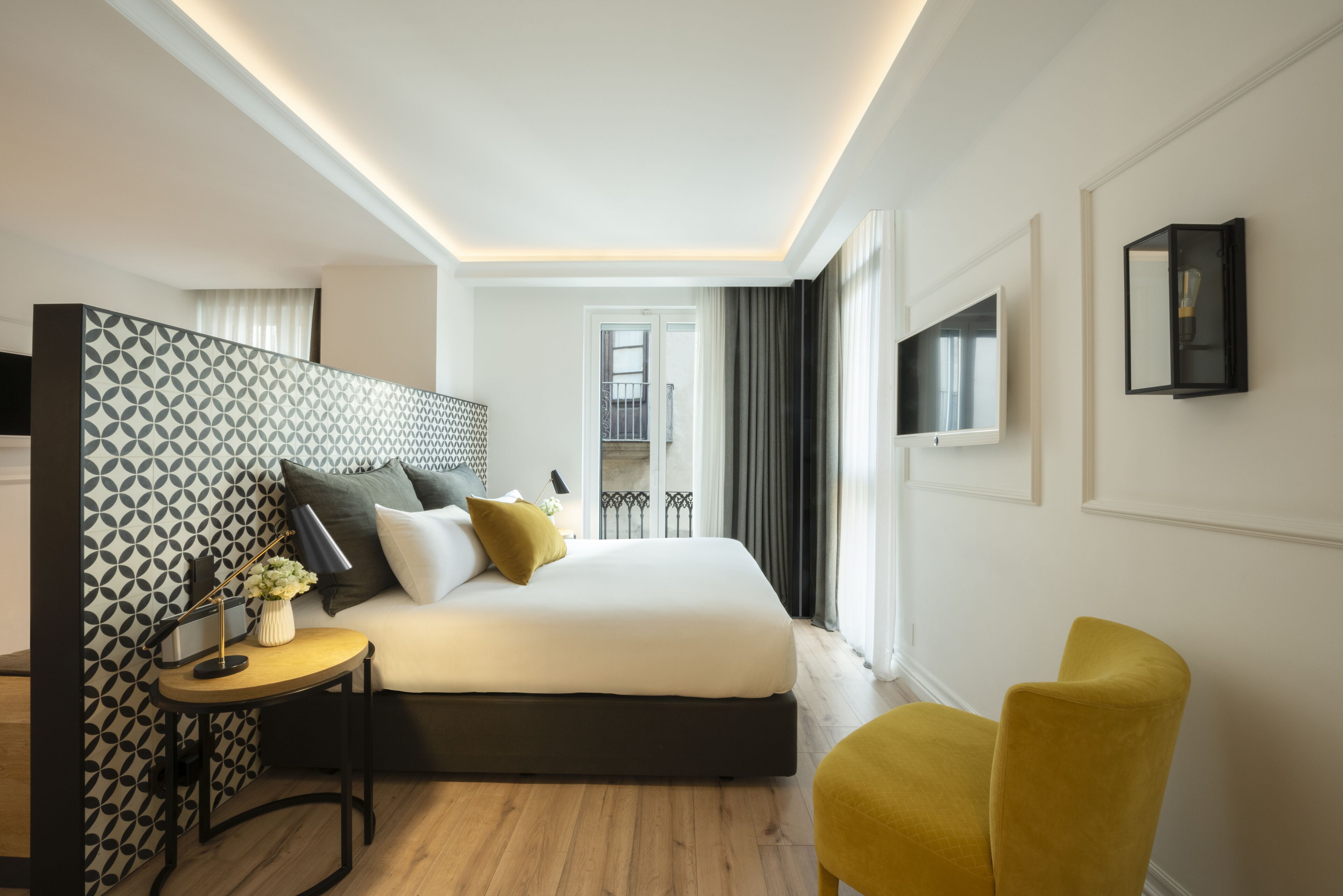 Una habitación de hotel moderna con una cama grande, una mesita auxiliar pequeña, un sillón amarillo y vistas al balcón.