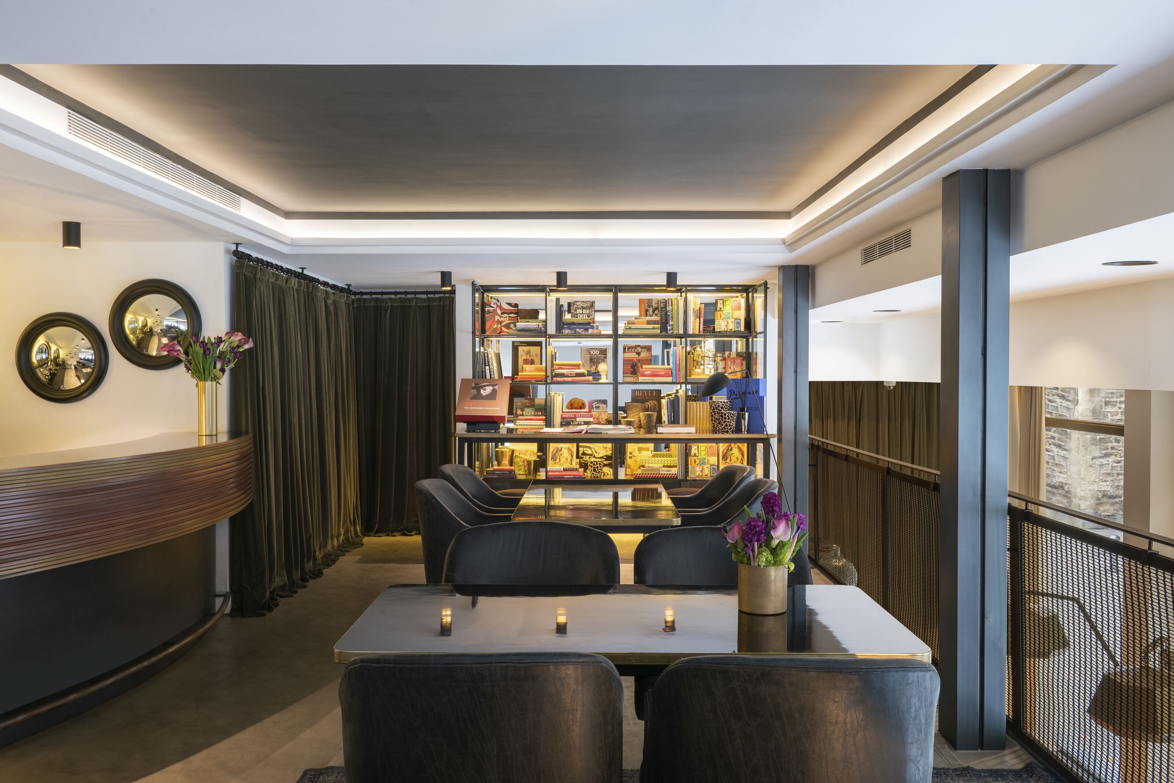 Un moderno vestíbulo de hotel con mostrador de recepción, zona de estar y una estantería llena de libros y objetos decorativos.