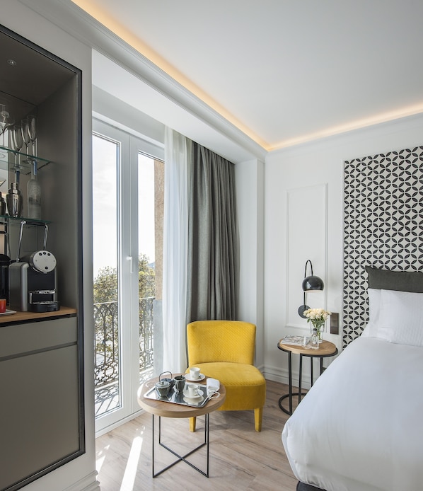 Une chambre d’hôtel moderne avec un grand lit, un fauteuil jaune, une petite table ronde avec un plateau, une machine à café et une vue sur l’extérieur.