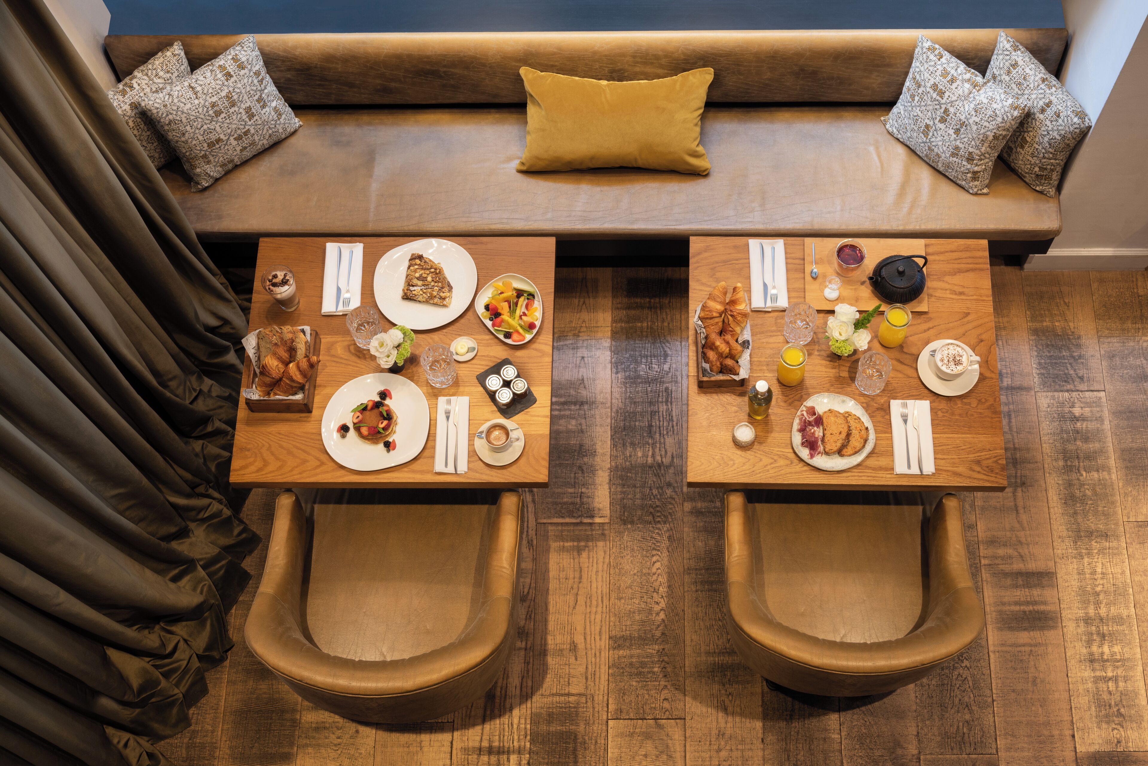 A dining area with two wooden tables, each with a plate of food, a glass of drink, and a cup. There are two brown leather chairs and a sofa with patterned cushions.