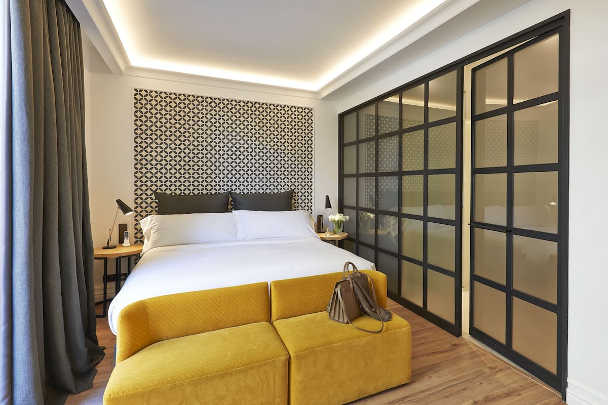 A hotel room with a bed, a yellow ottoman, a nightstand, a lamp, and a large window with frosted glass panels.