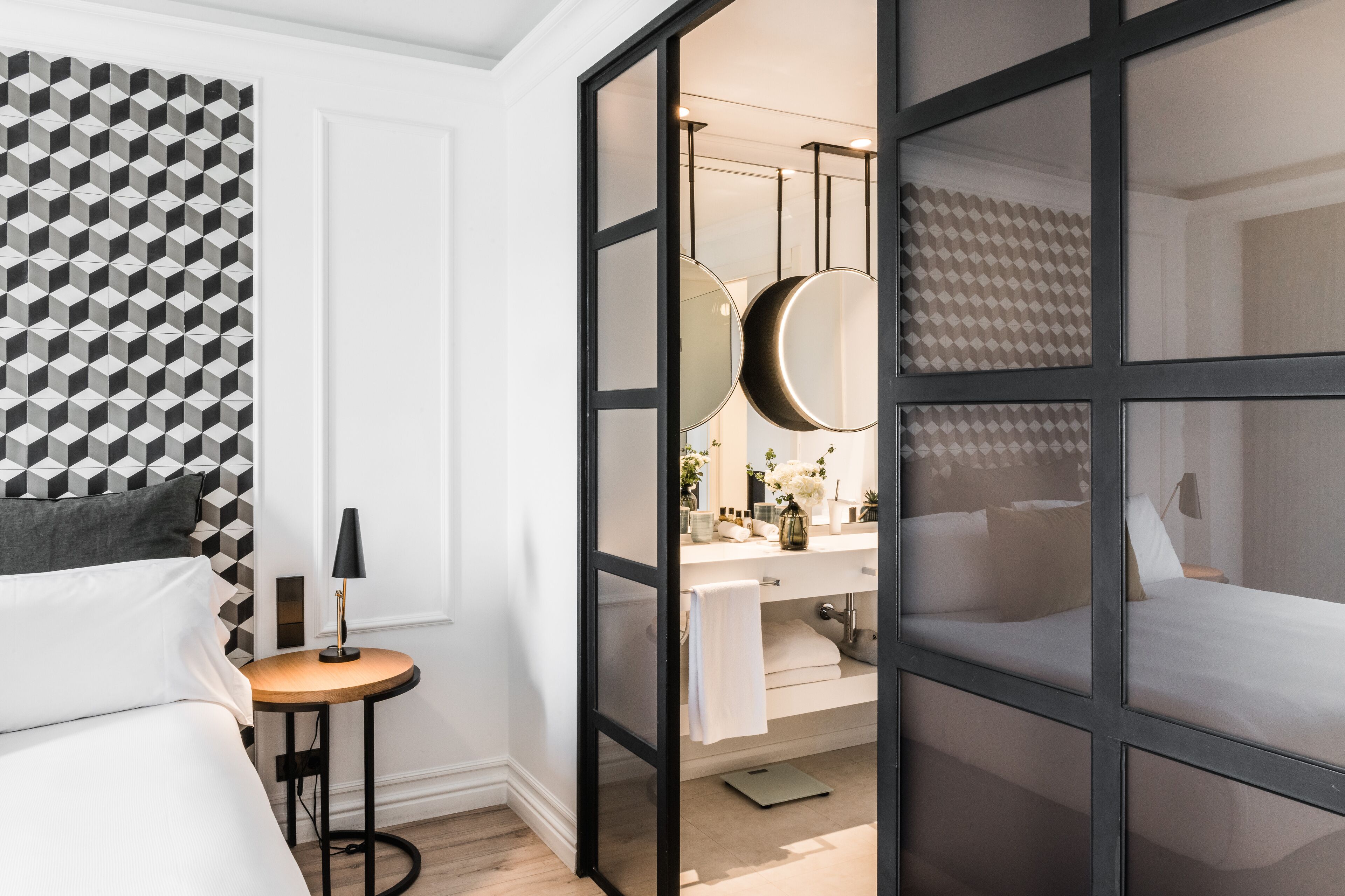 Una habitación de hotel moderna con un papel tapiz de diseño geométrico, una mesita redonda con lámpara, una cama con ropa blanca, un baño con espejo y lavabo, y una puerta corredera que da a otra habitación.