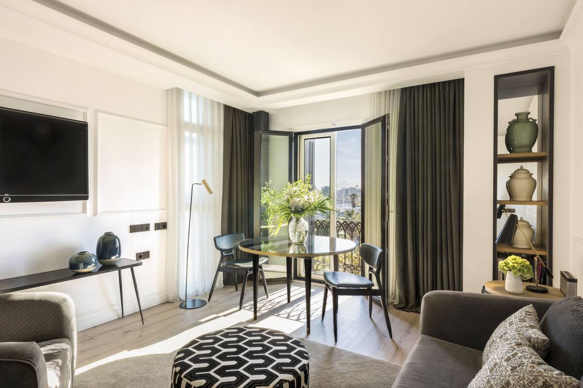 A modern living room with a round glass table, two chairs, a sofa, a TV mounted on the wall, and a balcony with a view of the city.