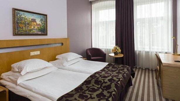 A hotel room with a double bed, a desk with a chair, a small table with a vase of flowers, and a painting on the wall. - Hotel London Tartu (Tartu)