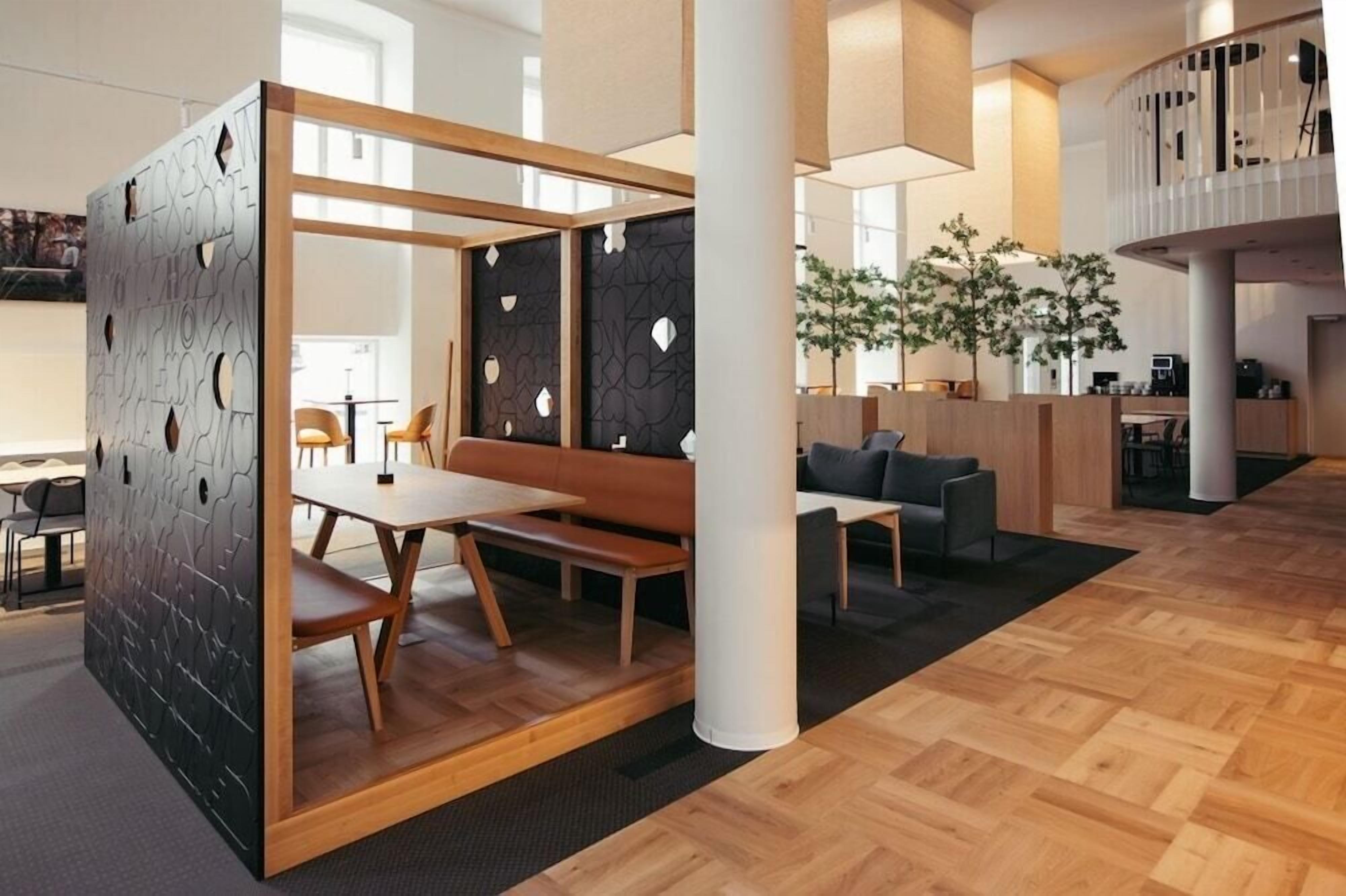 Et moderne interiør med trægulv, et siddeområde med bænk og stole, samt en rumdeler med geometrisk mønster.