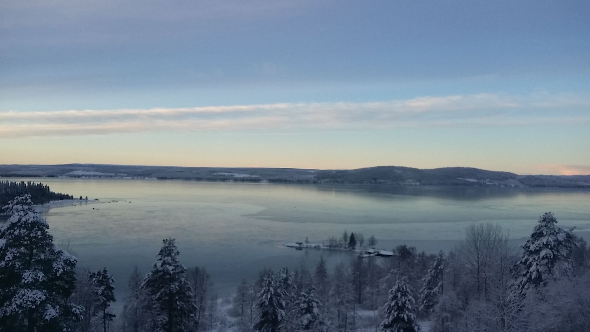 A panoramic view of a lake surrounded by snow-covered trees and distant hills.