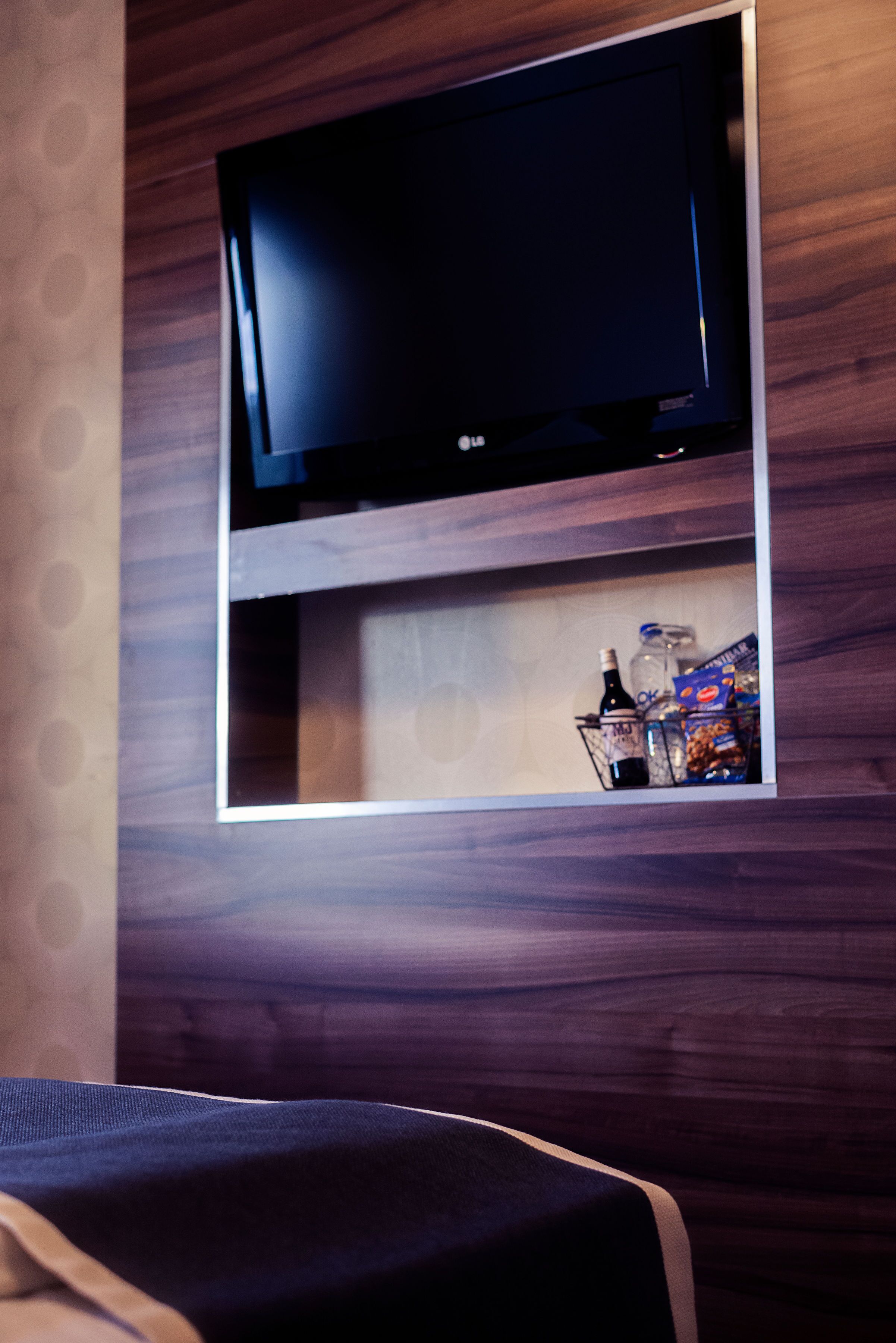 A television mounted on a wooden wall with a shelf below it containing a bottle, a box, and some snacks.