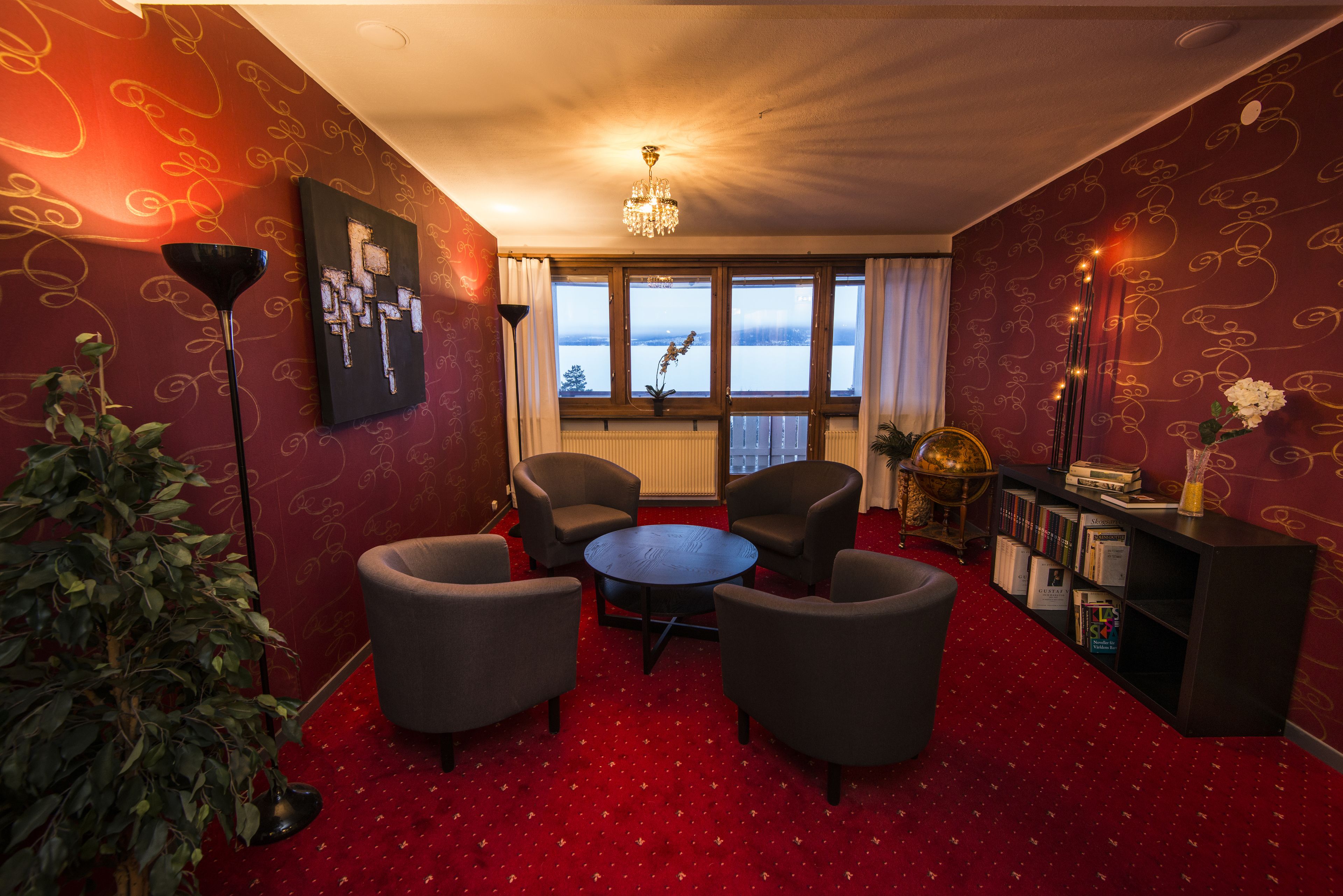 Ein Zimmer mit rotem Teppich, einem runden Tisch, vier Sesseln, einem Bücherregal, einer Topfpflanze, einem Gemälde und Blick auf einen See durch große Fenster.