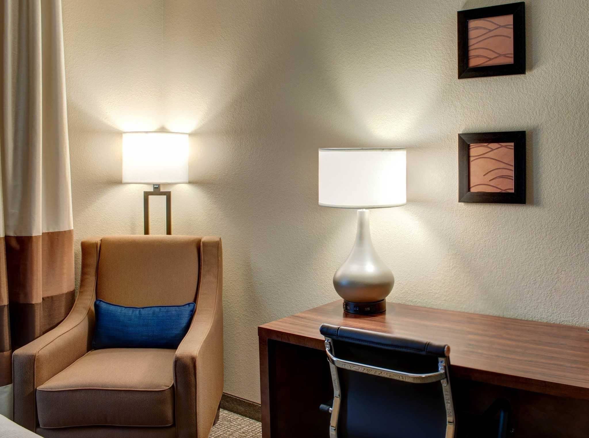 Une chambre d’hôtel avec un fauteuil beige, un bureau en bois et deux lampes fixées au mur.