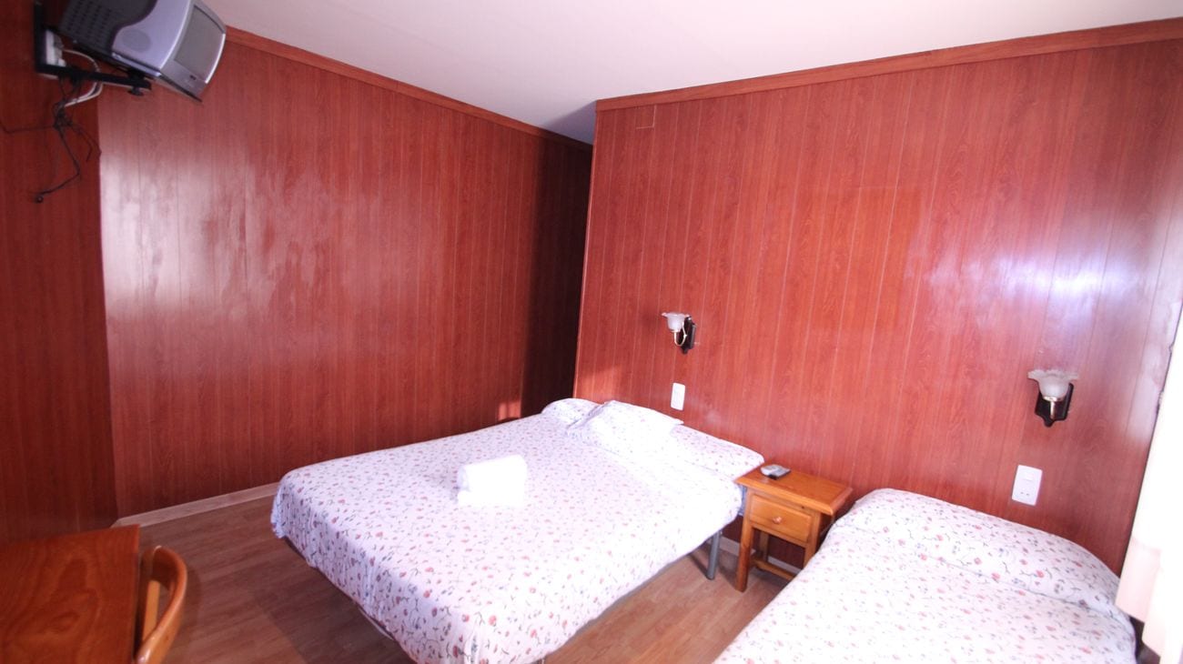 A small, compact room with two single beds, a wooden desk, a chair, and a television mounted on the wall.