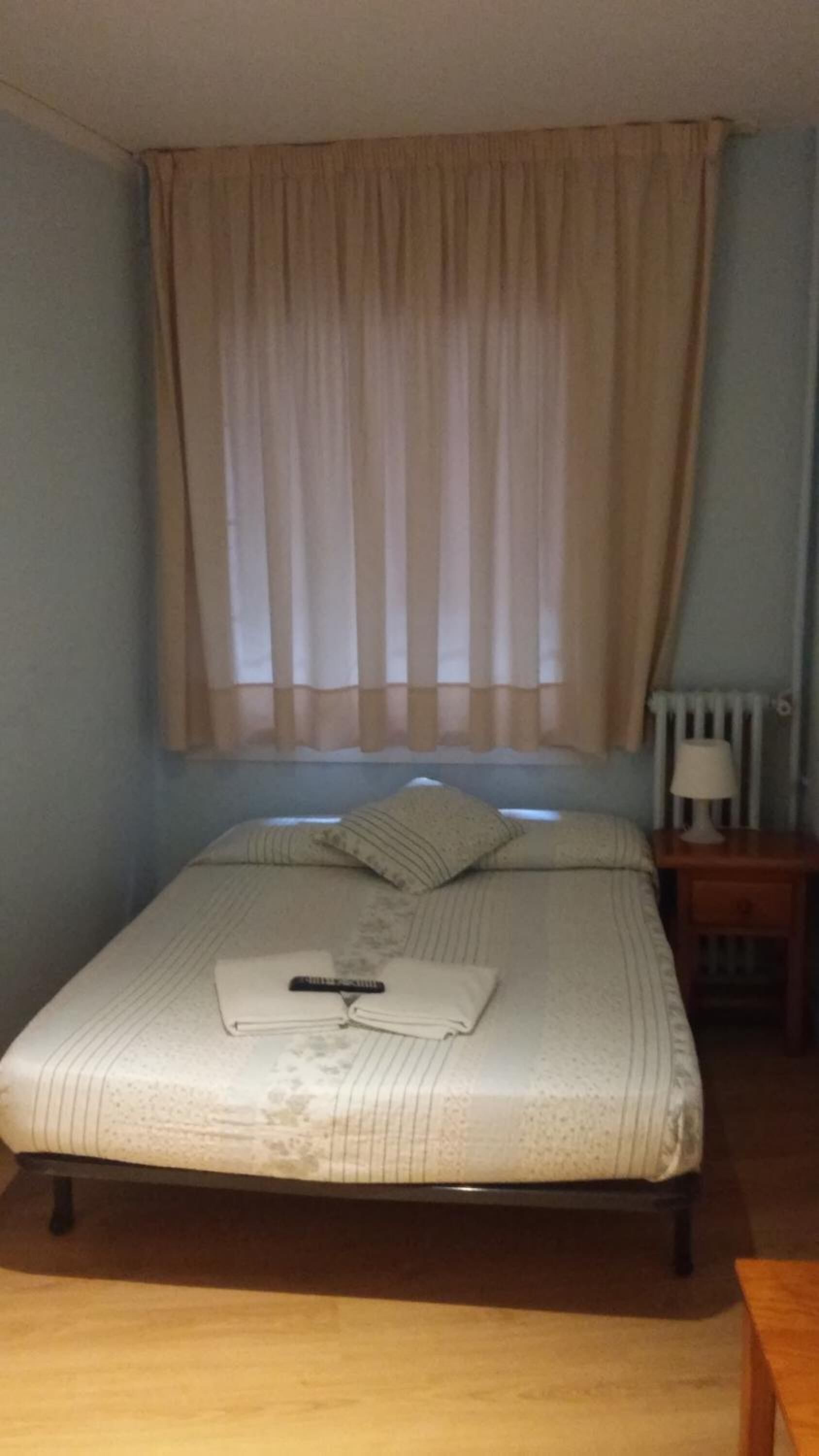 Una cama individual con manta y almohadas, una mesita de noche con lámpara, y una ventana con cortinas.