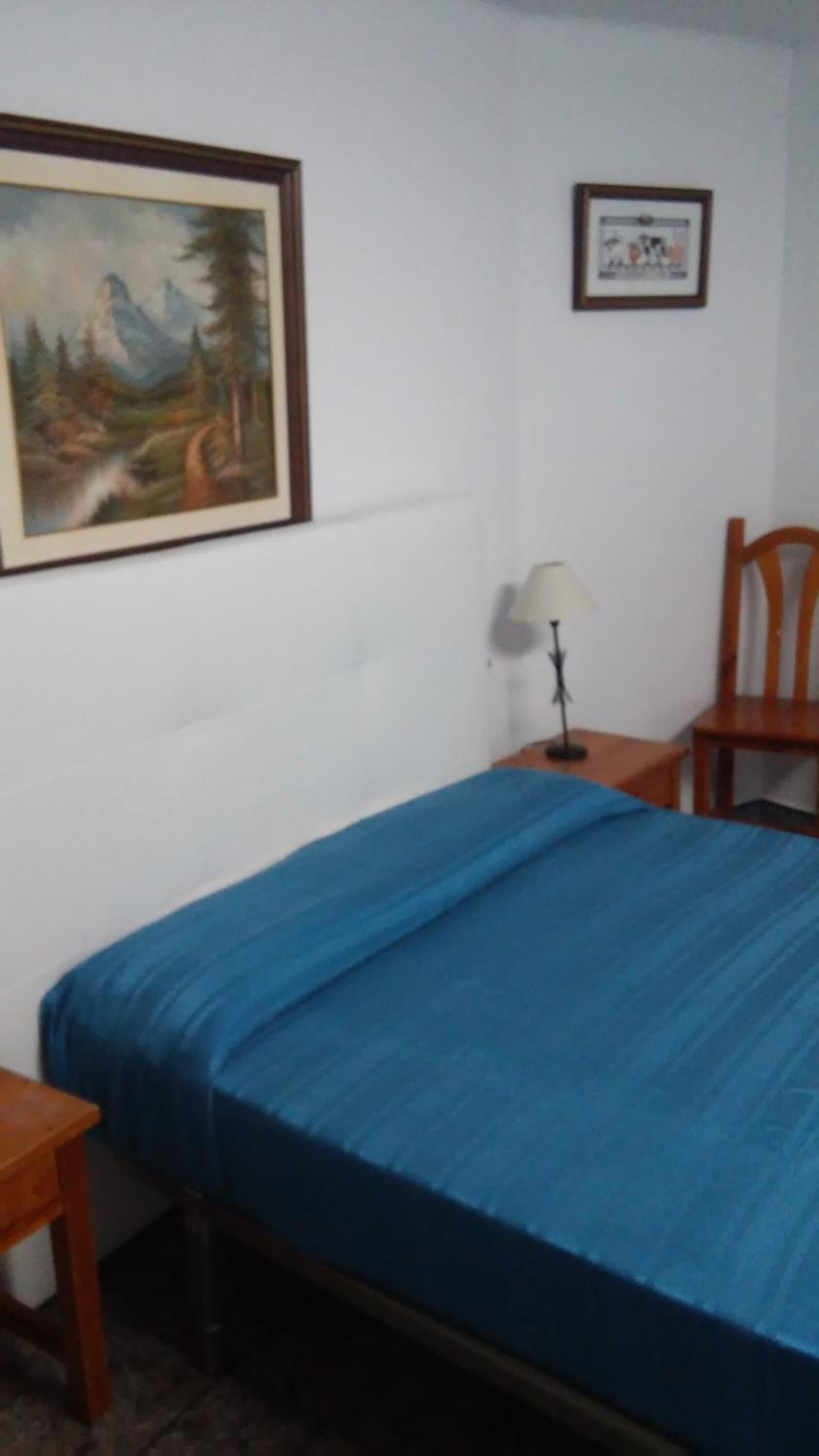 Una habitación con una cama, un escritorio, una silla, una lámpara y dos cuadros en la pared.