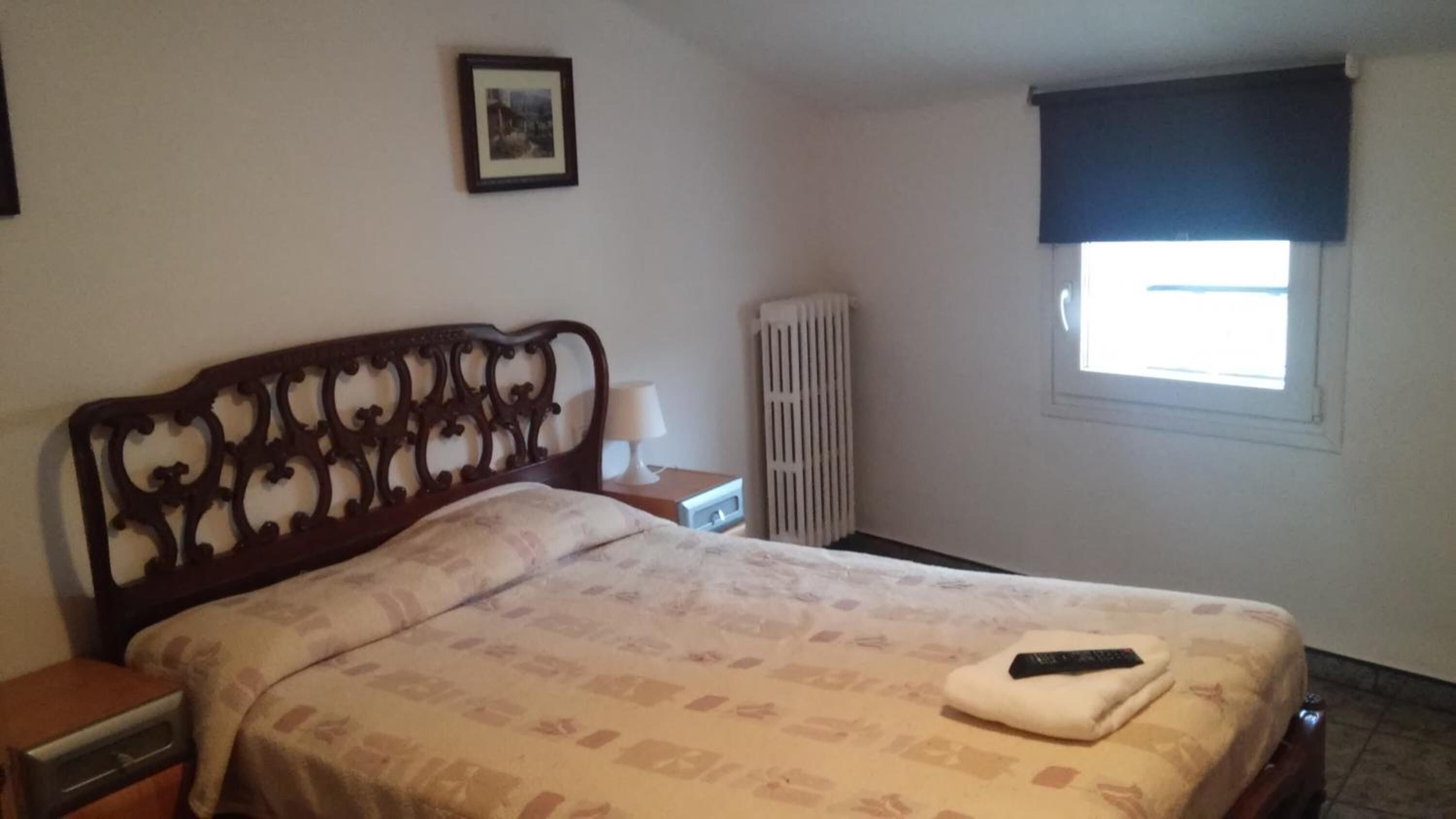A bedroom with a wooden headboard, a bed with a patterned bedspread, a bedside table with a lamp, a radiator, and a window with a dark blind.
