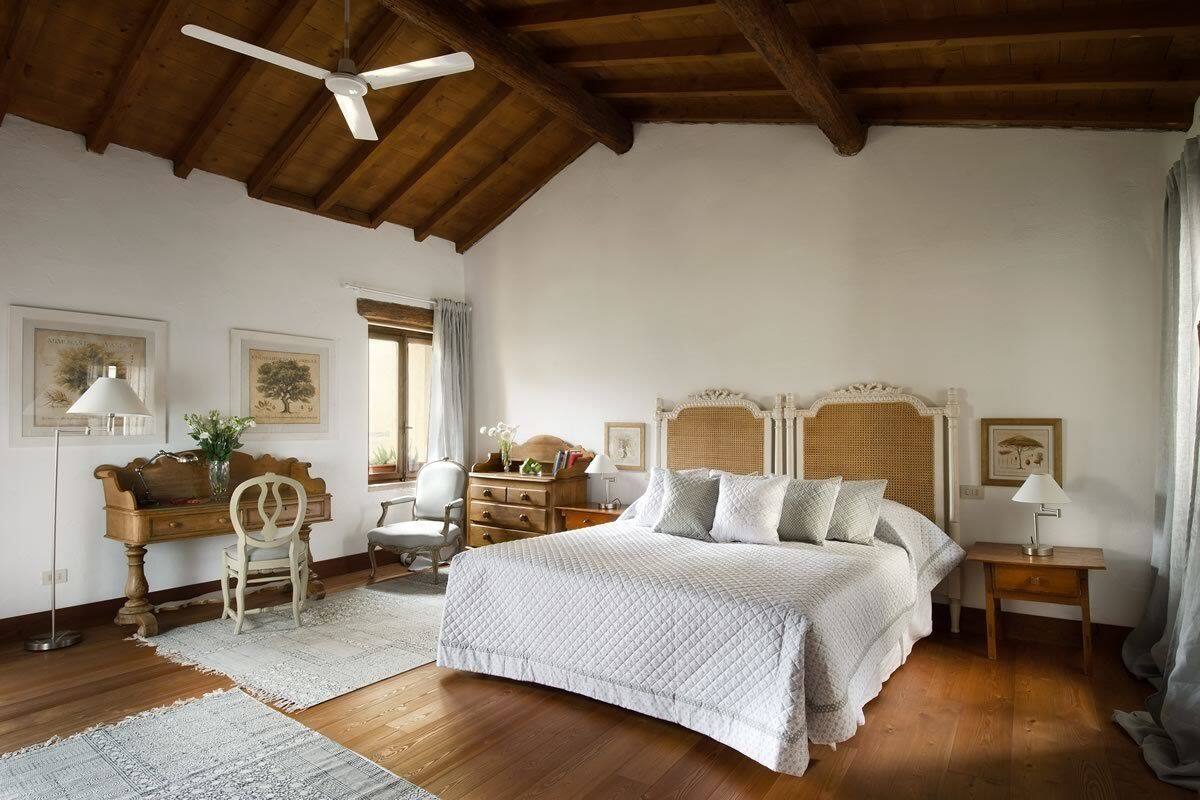 A bedroom with a wooden ceiling, a large bed with a headboard, a desk with a chair, and a window with curtains.