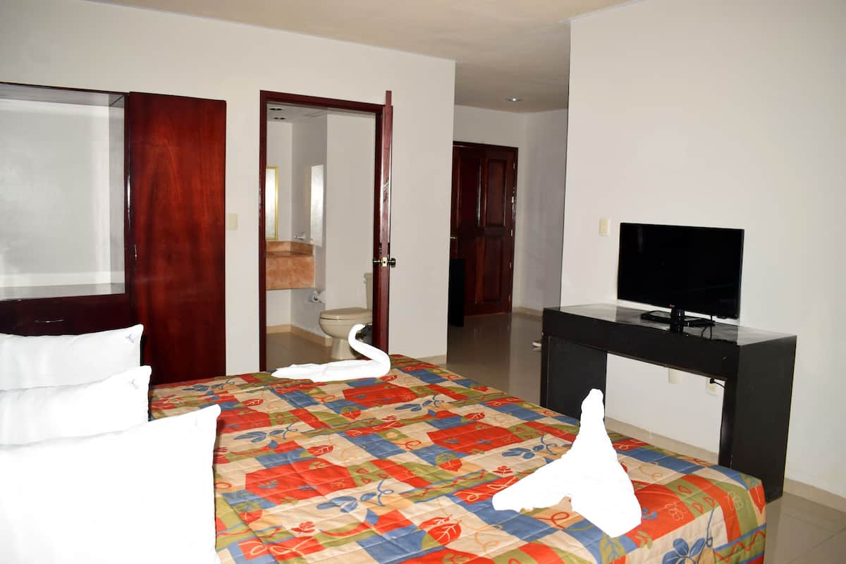 Quarto de hotel com uma cama, televisão, escrivaninha e banheiro visível por uma porta aberta.