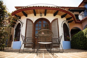 Un patio con mesa y sillas, un área cubierta con techo de tejas y un edificio con fachada y ventanas de arco.