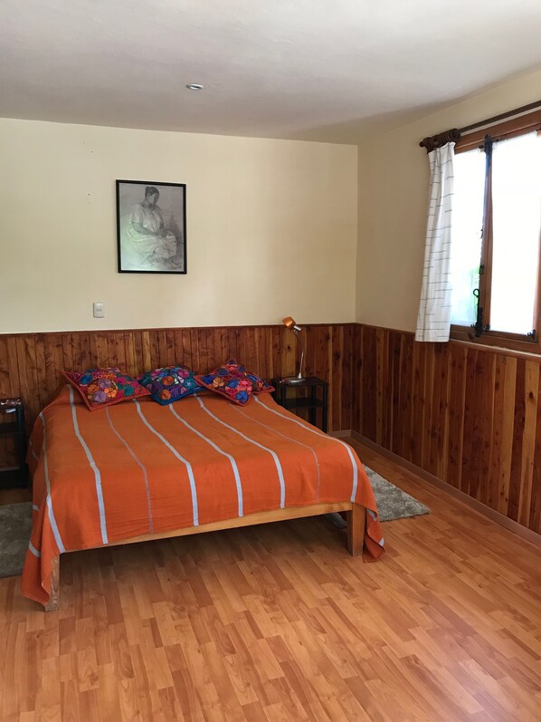 Un dormitorio con una pared de paneles de madera, una cama con colcha naranja, un cuadro enmarcado en la pared y una ventana con cortinas.