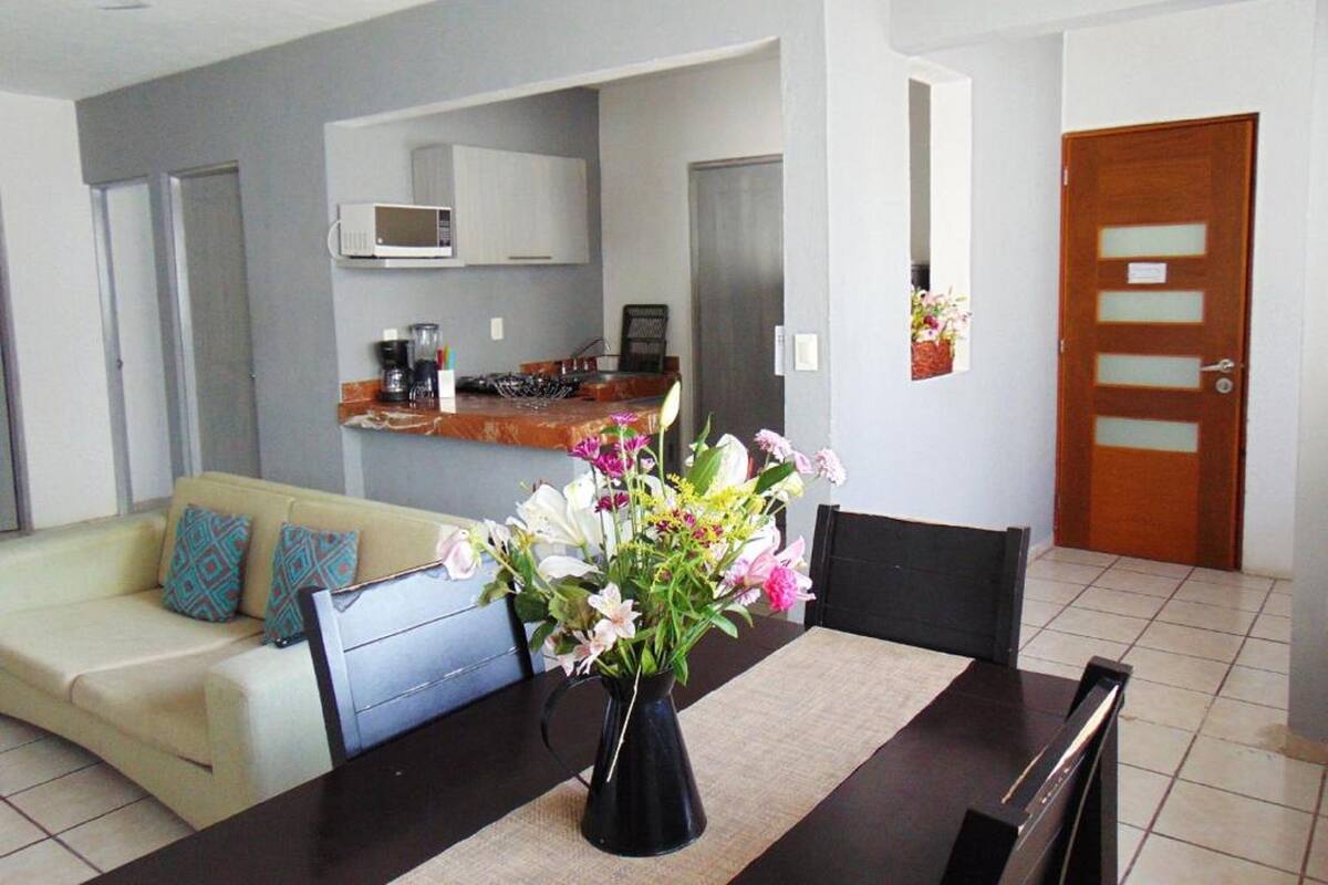A dining area with a table, chairs, and a vase of flowers. There is a kitchen in the background with a microwave and a coffee maker.
