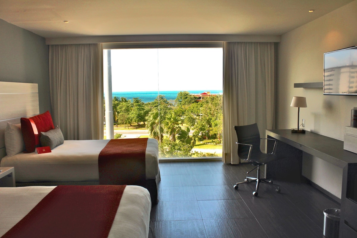 A hotel room with two beds, a desk, a chair, and a large window offering a view of the ocean and greenery.