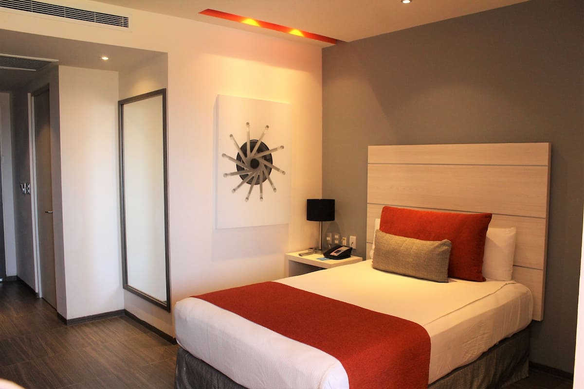 A hotel room with a large bed, a bedside table with a lamp, a wall clock, and a door to another room.