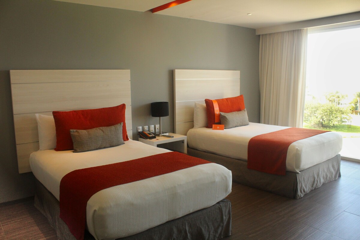Habitación de hotel con dos camas, cada una con almohada roja y gris, una mesita de noche con lámpara y un ventanal con cortinas.