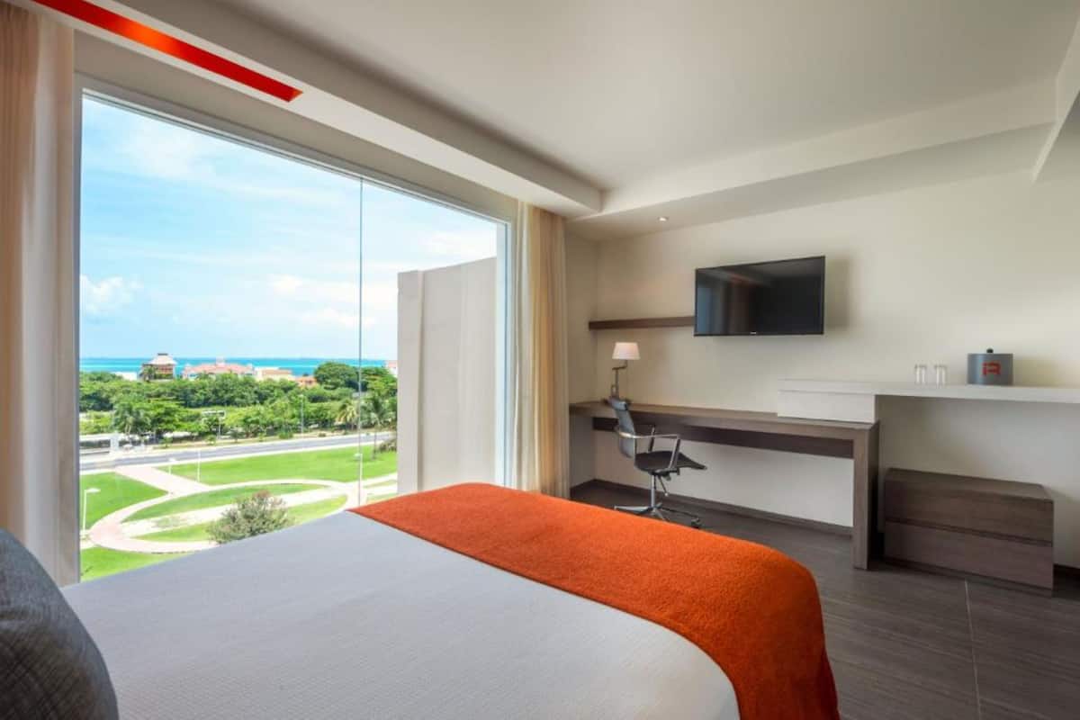 A hotel room with a large bed, a desk with a chair, a flat-screen TV, and a view of a park and buildings.