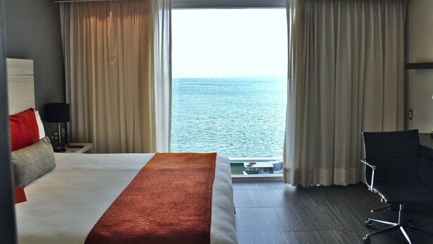 A hotel room with a large bed, a desk, a chair, and a view of the sea through the windows. - Real Inn Cancun (Cancun)