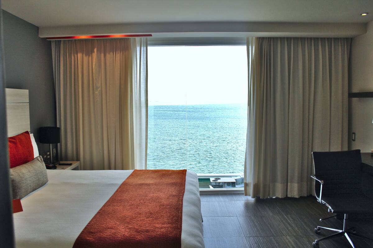 A hotel room with a large bed, a desk, a chair, and a view of the sea through the windows.