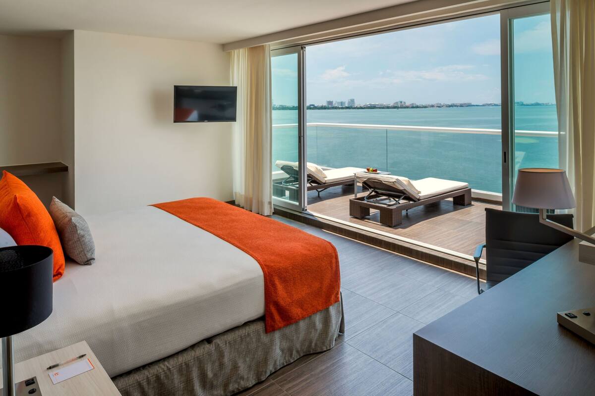A modern hotel room with a large bed, a desk, a chair, and a view of the sea.