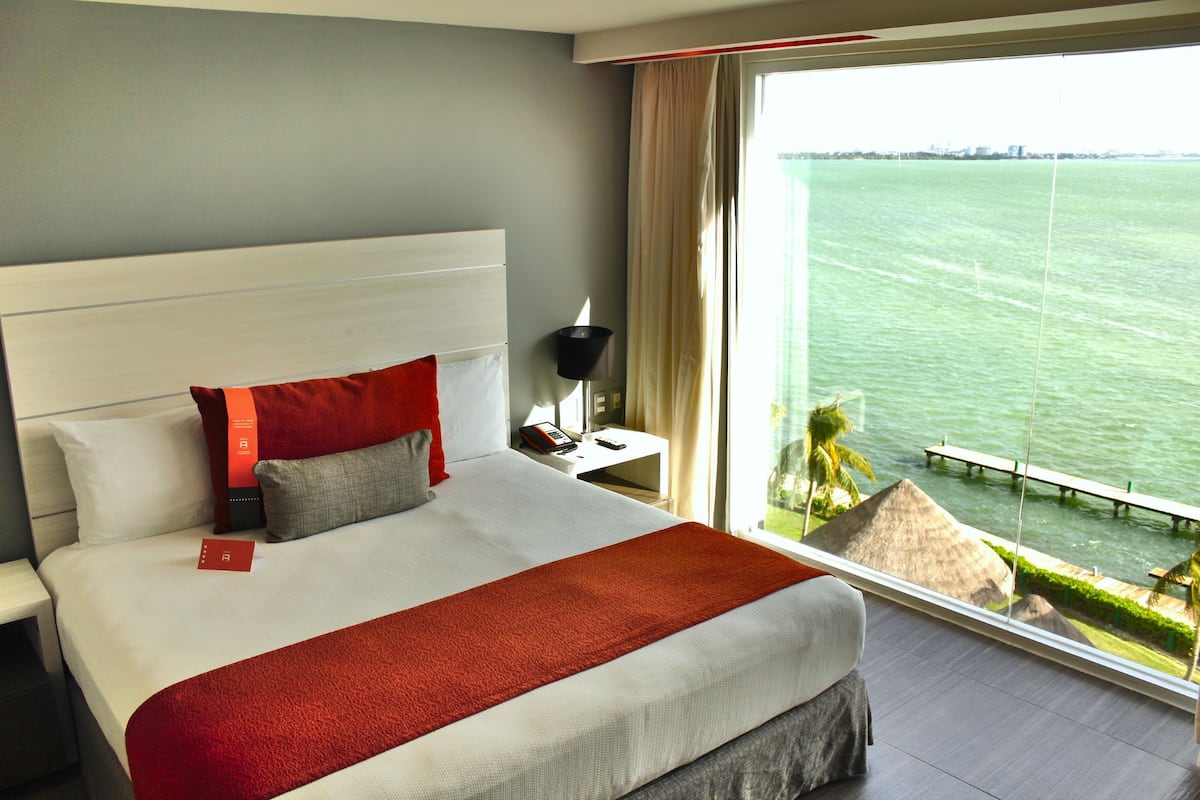 A hotel room with a large bed, a nightstand, a lamp, and a view of the sea.