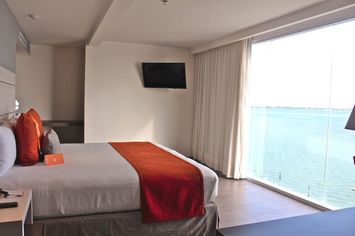 A hotel room with a bed, a TV mounted on the wall, a window with a view of the sea, and a table with a remote control.