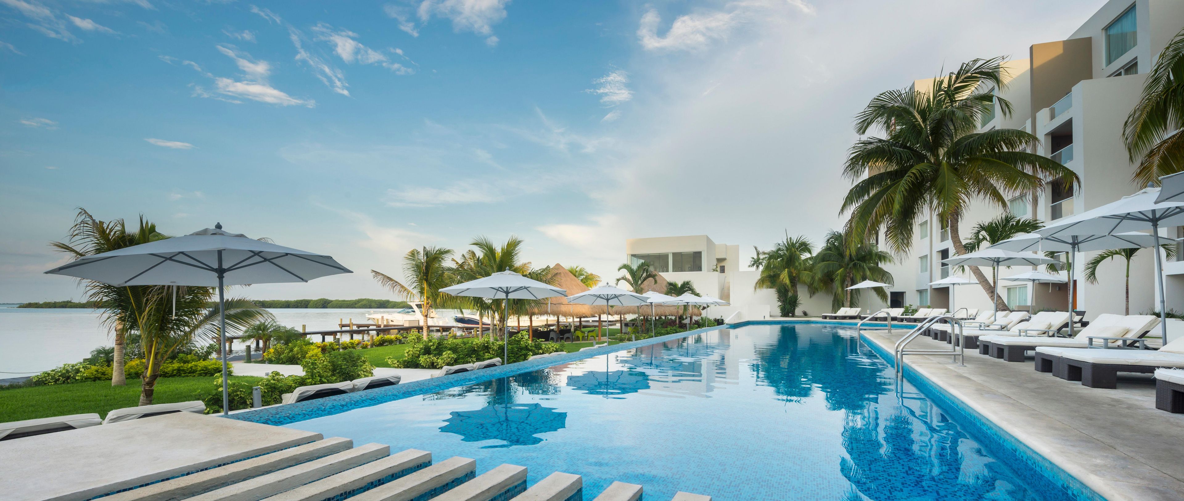 A swimming pool with lounge chairs and umbrellas, surrounded by palm trees and a view of the water.